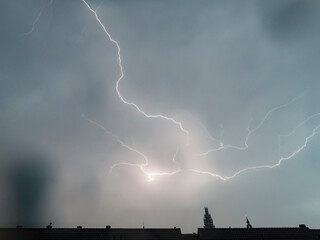 Powerful lightning strikes illuminate a gray storm sky.  Rooftops are silhouetted below.