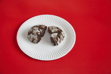 A broken chocolate cookie with cracks lying on a white paper plate against a red background, close-up, homemade baking, chocolate cookies, treats on a plate