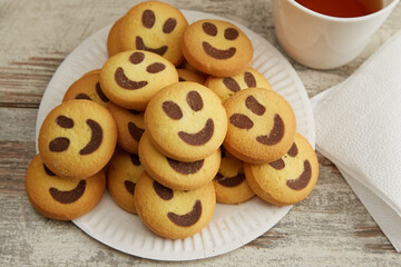Smiley cookies arranged in a pile on a white paper plate on a wooden table next to a cup of tea and a napkin, close-up, sweets with tea, good breakfast culinary recipe.