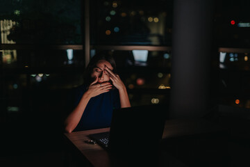 A stressed businesswoman sits alone in a dark office, working on her laptop. The dim light and her...