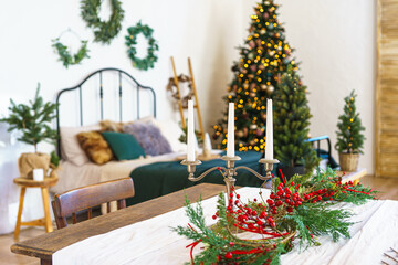 Three candles in candelabra on table surrounded by red berries and greenery on Christmas tree glows with warm lights background. Festive bedroom interior decorated for winter holidays