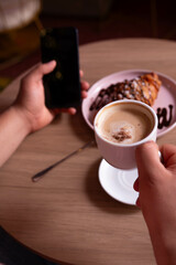 Close-up of a cup of cappuccino coffee. Man holding his smartphone while drinking coffee