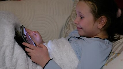 A 9-year-old Caucasian girl is lying on a sofa and playing computer games on a tablet. Shows joy