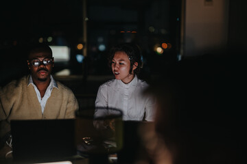 A diverse group of coworkers collaborate in a dimly lit office, focused on meeting deadlines and strategizing projects during a late-night work session, illustrating teamwork and dedication.