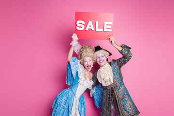 Excited couple dressed in elaborate baroque costumes posing with a sale sign against a vibrant pink background