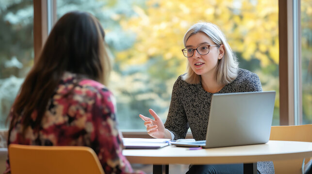 University professor guiding graduate student, reviewing materials together on round table with shared laptop during consultation