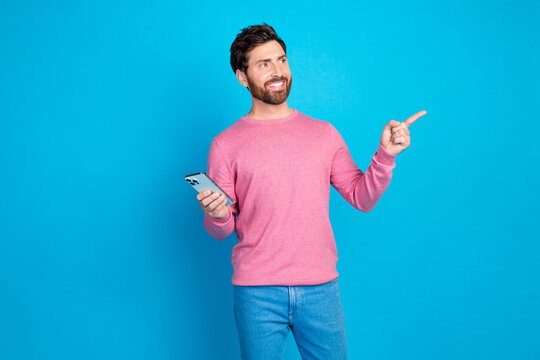 Confident bearded man in casual attire holding smartphone and pointing, standing against vibrant blue backdrop