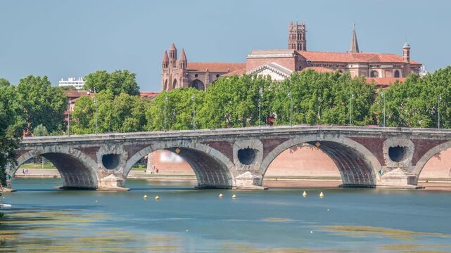 Garonne River and Pont Neuf timelapse with Basilica of Our Lady of the Daurade in downtown Toulouse, France. Renaissance arch bridge reflects in the water under a blue sky. Waterfront with green trees