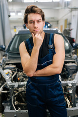 Vertical portrait of pensive mechanic male in jumpsuit serious looking at camera not knowing what disrepair during maintenance car in workshop. Doubtful repairman trying to solve problem in garage