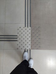 Feet in white sneakers in front of tactile tiles for visually impaired people on the floor indoors.