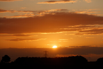 Sunset sky with orange and gold hues, clouds, and silhouetted trees.