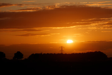 Fiery orange sunset over a silhouetted landscape with power lines and trees.  Golden light bathes the sky.