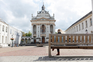 Obraz premium Person sits on park bench in front of ornate white church. Sunny day, paved square.