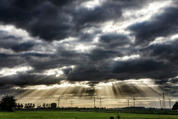 Dramatic sky with rays of sunlight piercing dark clouds over a field with wind turbines and power...