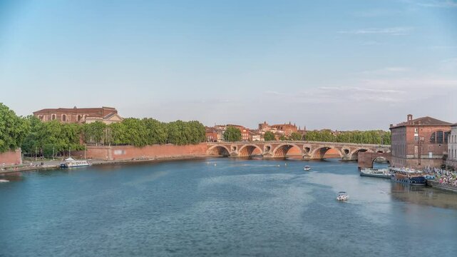 Garonne River and Pont Neuf timelapse with Port de la Daurade in downtown Toulouse, France. This Renaissance arch bridge reflects in the water under a blue sky with clouds. Waterfront with green trees