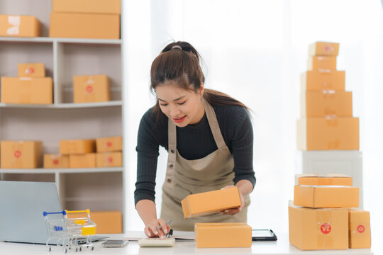 Busy entrepreneur: A young woman works diligently in her home office, surrounded by stacks of cardboard boxes. Her focus on her business is evident as she carefully prepares a package for shipment. - Powered by Adobe
