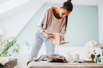 A young student is focused on organizing her study materials in a cozy, plant-filled room. She is flipping through a notebook, emphasizing preparation and concentration.