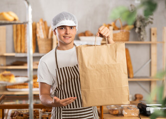 Portrait of positive young baker with paper bag of fresh baked goods in the interior of a private...