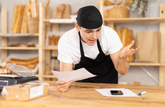 Young bakery owner in black uniform leaning on wooden counter surrounded by baked goods, using phone while examining paperwork with puzzled, worried expression, uncovering problems with order details - Powered by Adobe