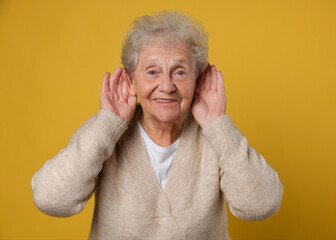 Senior woman showing hand to ear gesture on dark yellow background