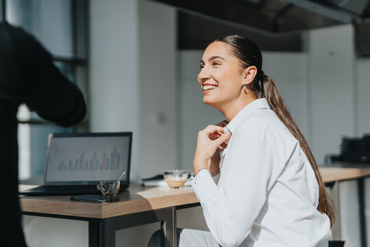 Business employees engaged in a conversation in a modern office kitchen. A woman is smiling while another person gestures towards a laptop. Bright and open workspace fostering collaboration.