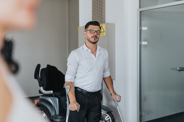 A young businessman using crutches stands confidently in a modern office environment. His...
