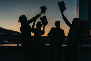 Obraz premium Silhouetted business people raising documents and celebrating success against a picturesque sunset. The group is outdoors, showcasing teamwork, achievement, and triumph in a professional setting.