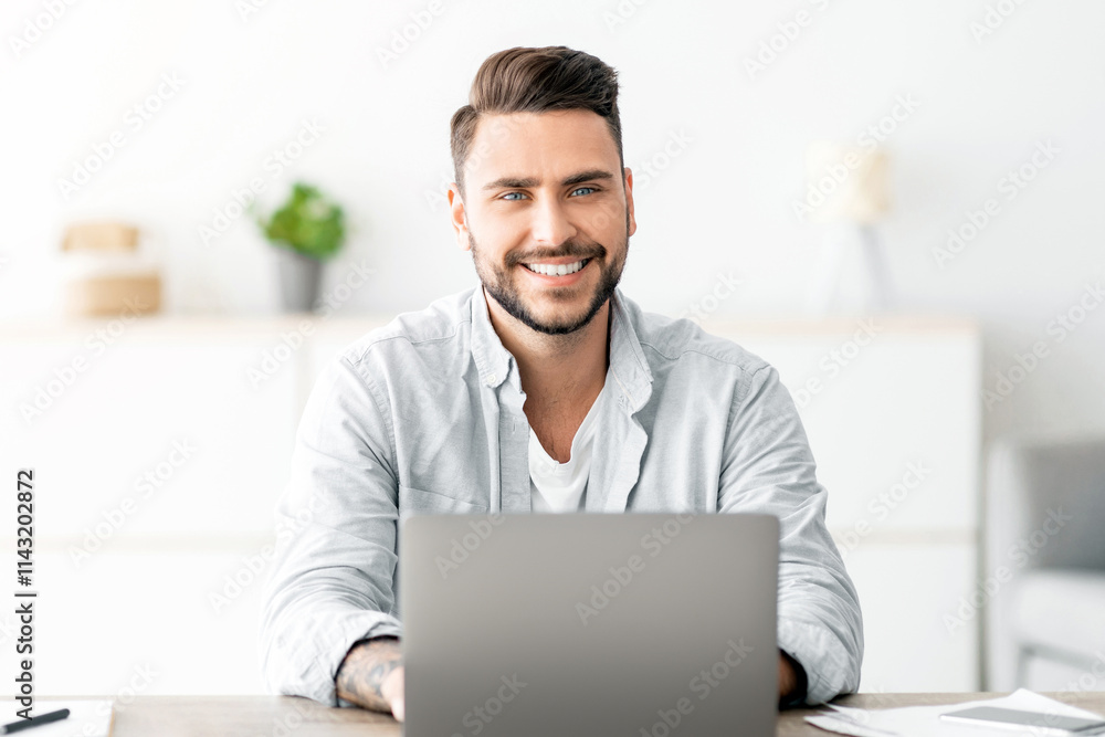 Poster successful freelancer. happy young man sitting at workplace with laptop and smiling to camera while 