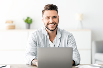 Successful freelancer. Happy young man sitting at workplace with laptop and smiling to camera while working on computer at home office, copy space. Guy enjoying remote work and self-employment