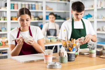 Concentrated young man and woman in aprons making handmade cups of row clay material in pottery workshop