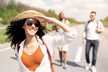 Cool millennial lady with multiracial friends hitchhiking on highway, traveling by autostop. Young...