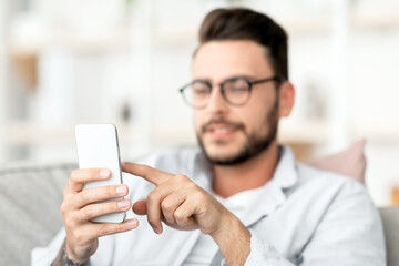 Handsome guy in eyeglasses using smatphone, touching screen while relaxing on couch at home, free space. Concentrated guy browsing internet or messaging with friends, sitting on sofa in living room