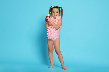 Cute little girl in swimsuit with drink on light blue background
