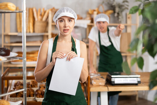 Young female bakery worker looking upset and uneasy while clutching piece of paper, while coworker angrily gesturing in background