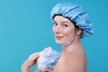 Woman in shower cap covered with soap foam holding mesh sponge on light blue background