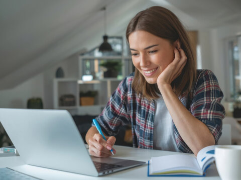 Mujer joven con una computadora portátil sentada en un escritorio. Ella está escribiendo notas mientras mira un seminario web. La mujer está estudiando en línea, aprendiendo clases web o teniendo una 