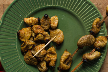 Overhead view of roasted sago worms on a green plate, top view of palm worms on wood background,...