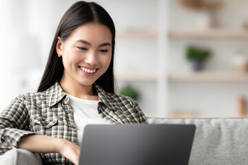 Cheerful asian woman typing on laptop keyboard, home interior, copy space. Positive korean young lady chatting with guys on dating website, reclining on couch in living room, copy space