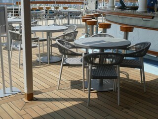 Empty round tables and wicker chairs stand on the wooden deck of a cruise ship, inviting passengers to relax and enjoy the sunshine