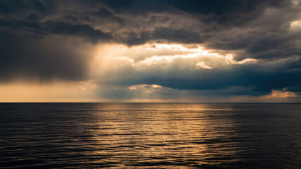 Obraz premium Dramatic sunset over calm waters, with dark clouds contrasting against golden rays of sunlight creating a tranquil yet powerful scene.