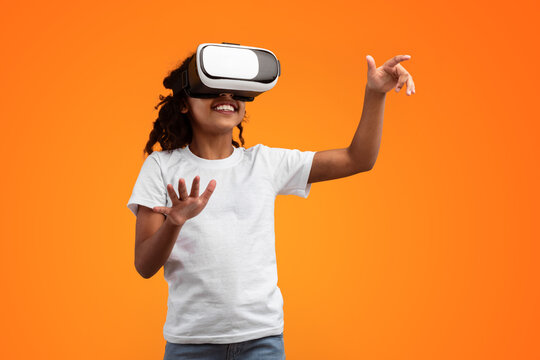 Virtual Reality Concept. Portrait of excited African American girl in VR headset touching air at yellow studio, copy space. Positive teen exploring artificial computer world or playing video game