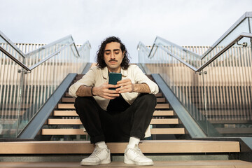 Casual modern man sitting on stairs using smartphone outdoors