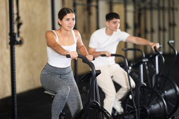 Positive girl riding exercise bike for cardio workout during cycling class at modern crossfit gym