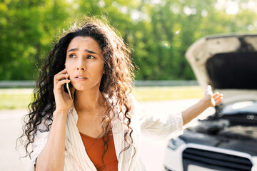 Road accident, car wreck, engine failure concept. Young woman with cellphone calling towing service on road, having phone conversation with mechanic near her broken auto on highway