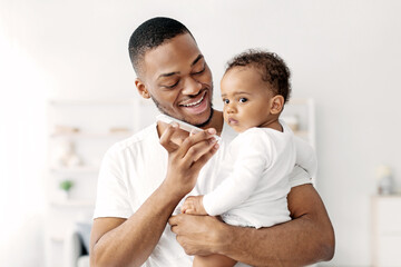 Obraz premium Modern Technologies And Babies. Black Father Holding Infant Child And Talking On Speakerphone At Home, Happy Young African American Man Joining Little Kid To Mobile Conversation, Free Space