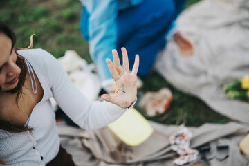 A woman shows her hand with colorful paint on her fingers, surrounded by picnic items. The outdoor setting captures a creative and relaxed atmosphere.