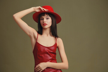 Young woman in a red hat and leather dress posing confidently against a green background, showcasing a bold and stylish fashion statement