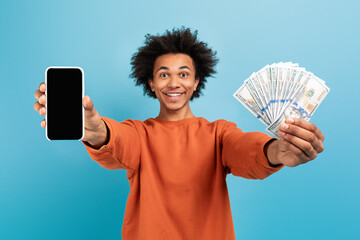 A young African American man smiles widely while holding a smartphone in one hand and a bundle of...