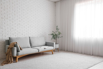 Stylish grey sofa with houseplant near white brick wall in room