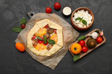 Baking paper with uncooked tomato galette and ingredients on black background